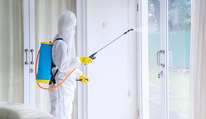 A remediation specialist in a full white biohazard suit and yellow gloves using a pressurized sprayer to disinfect high-touch surfaces in a bright residential living area