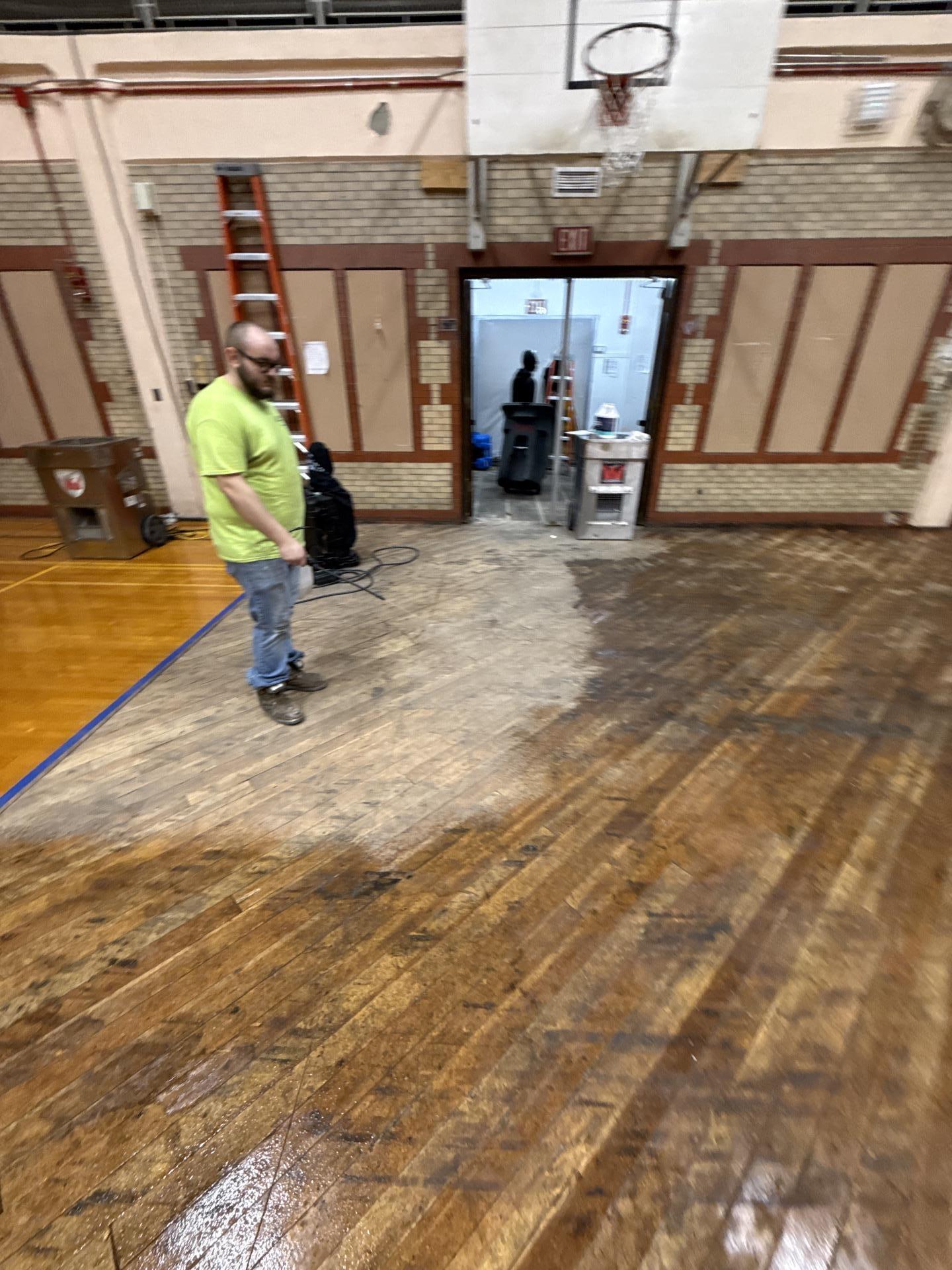 A restoration technician inspects a partially removed and water-damaged hardwood floor in a gymnasium, with basketball hoop and exit sign in the background.