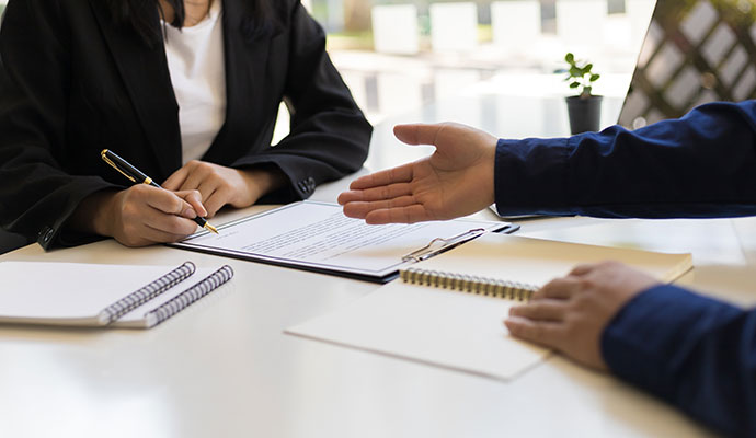 A person in a suit signing a document on a clipboard, sitting across from another person at a desk